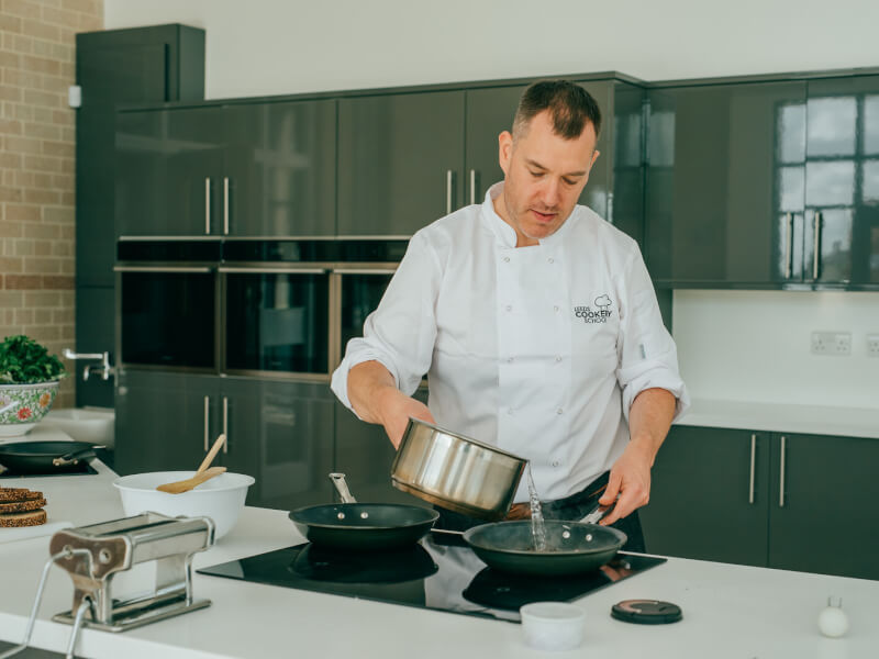 A man wearing chefs whites stands at the hob in a kitchen, pouring water from a saucepan into a frying pan